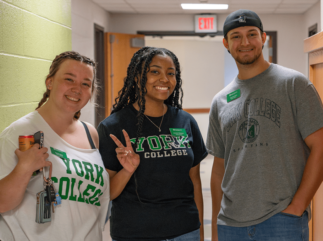 Three people wearing York College apparel smile at the camera in a residence hall setting.
