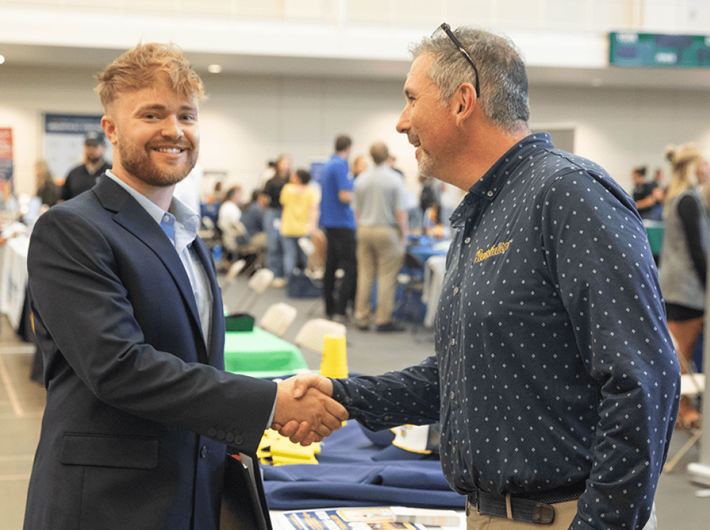 A student in business attire shakes hands with an employer at a career event.