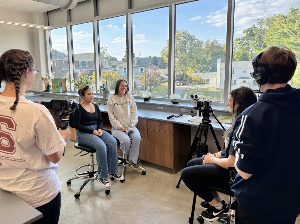 Two students sit facing a camera in a classroom setting. Two other students are behind the camera.