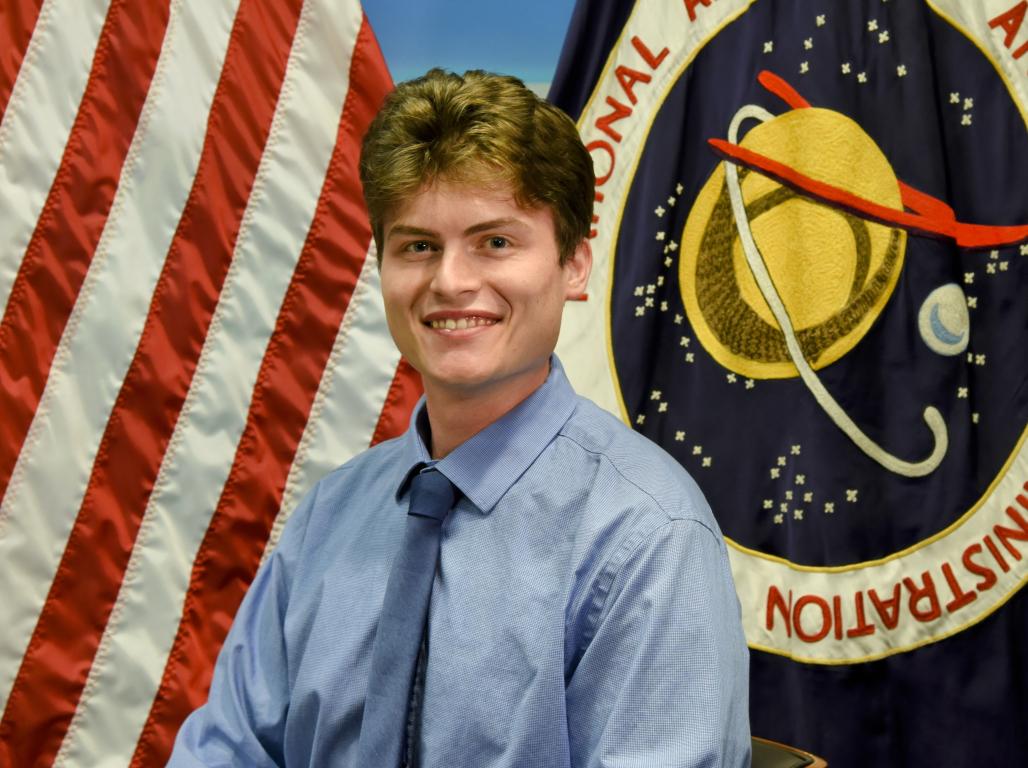 A professional headshot of Josh Polancheck with an American flag and NASA flag behind him