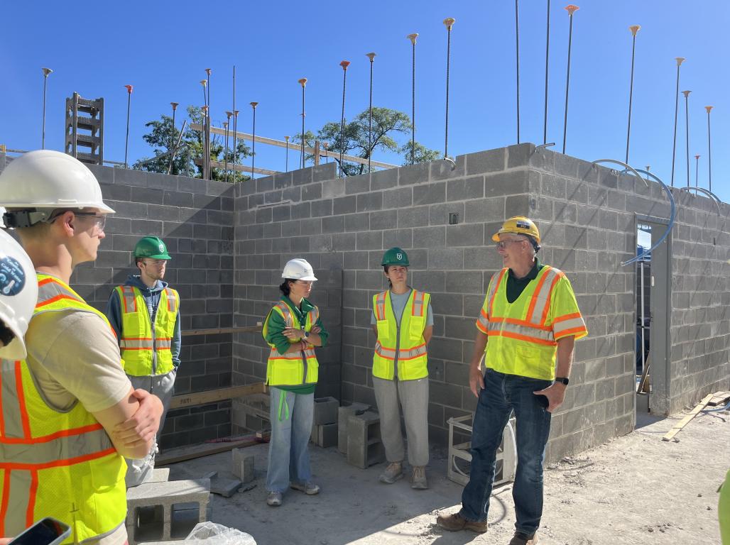 A group of engineering students listen attentively while touring a construction site, all are dressed in reflective vests and hard hats.