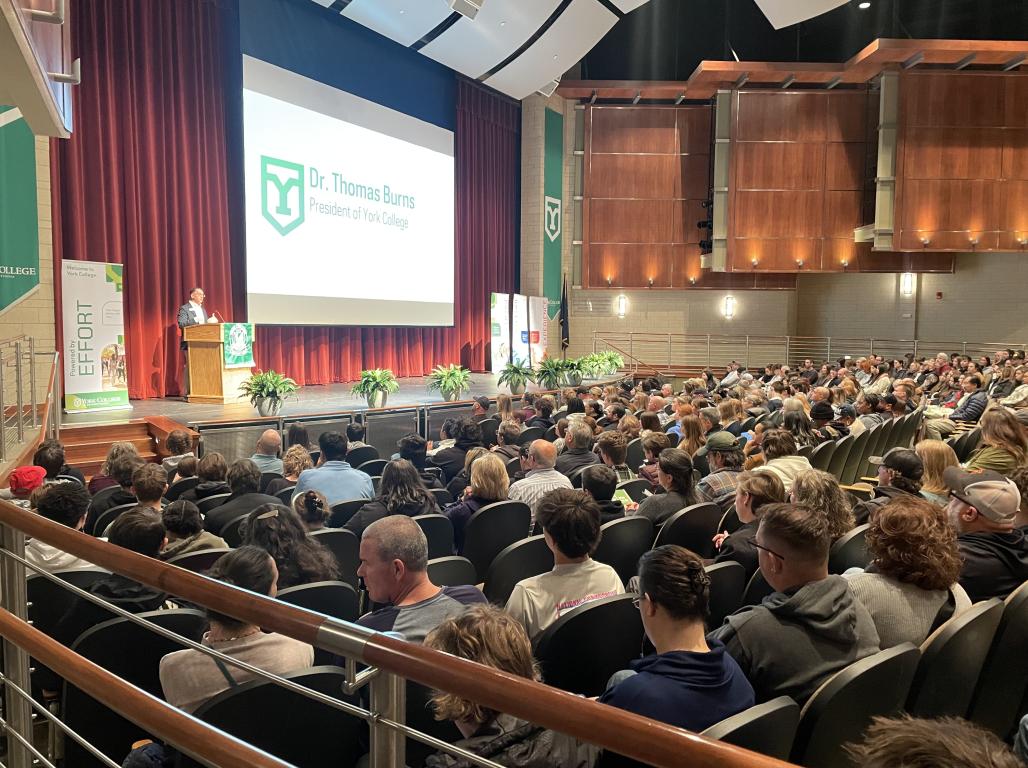 An auditorium filled with people listening to a speaker behind a podium.