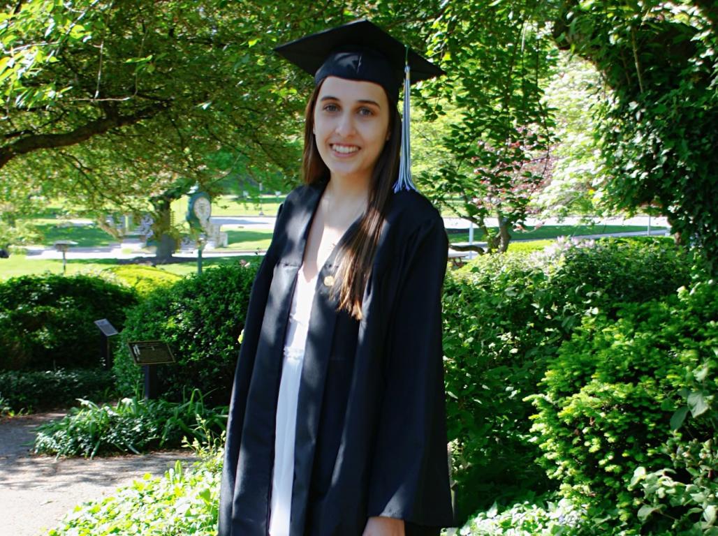 College graduate in regalia smiles at the camera on a sunny spring day.