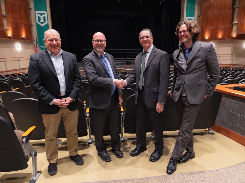 Four people in business suits smiling inside a theater, two are shaking hands.