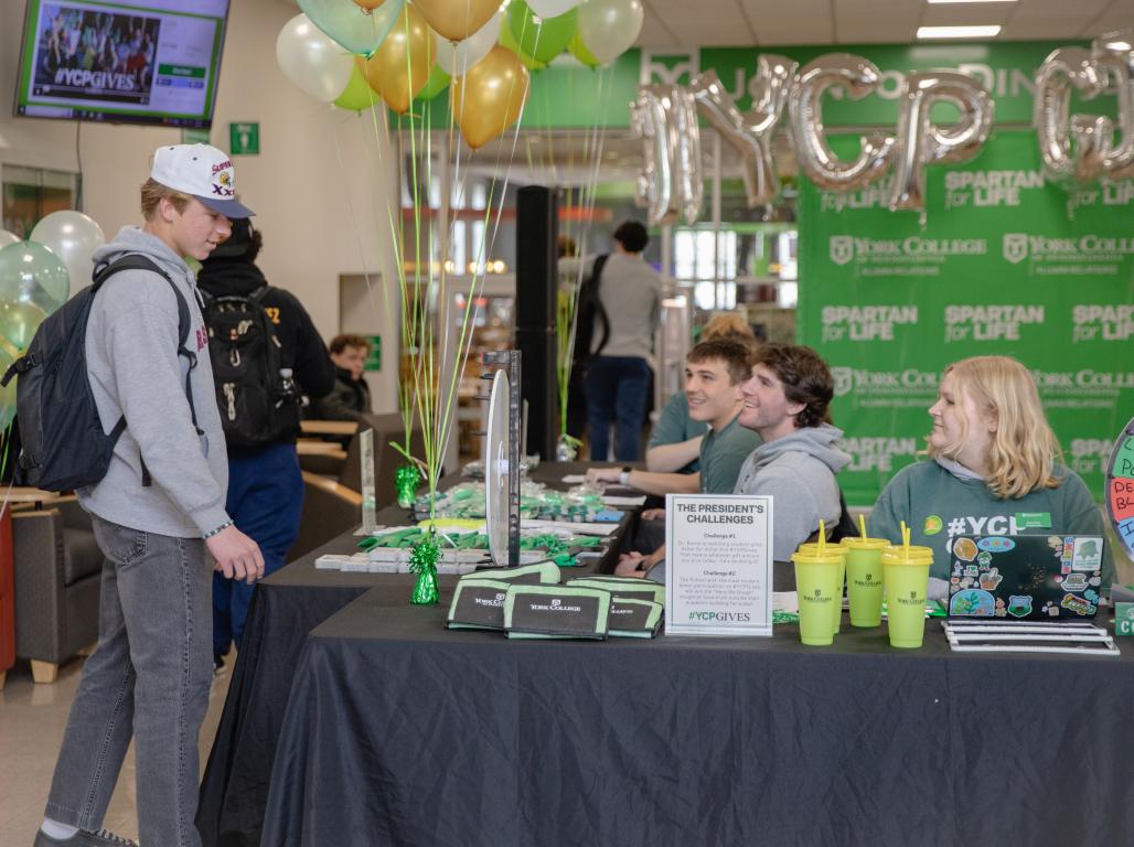 A student wearing casual clothes and a backpack speaking with other students who are seated behind a table covered in a black tablecloth with various promotional items on display. Balloons and a green backdrop reading "YCPGives" are visible behind them.