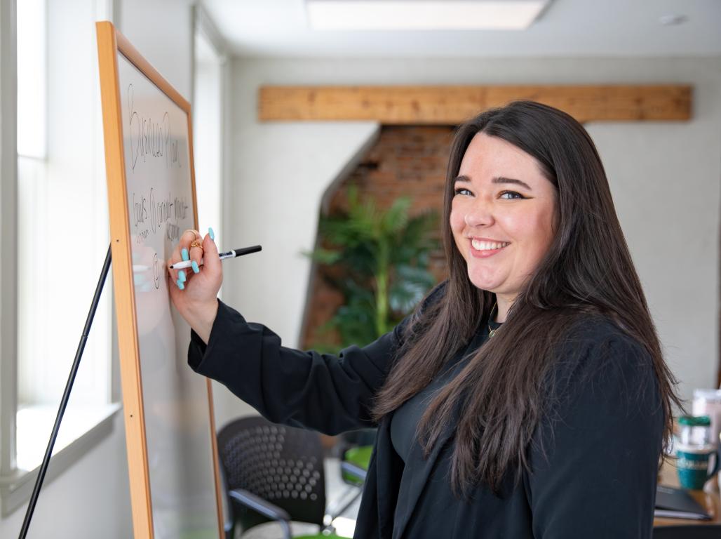 An adult student uses a dry erase marker on a white board. Her head is turned to the camera and she is smiling.