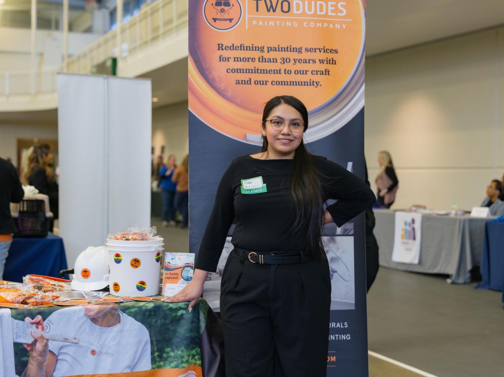 Person wearing black with a name tag reading "Cristal" and a green "Alumni" sticker beneath it stands in front of a banner for "Two Dudes Painting Company" at a professional networking event.