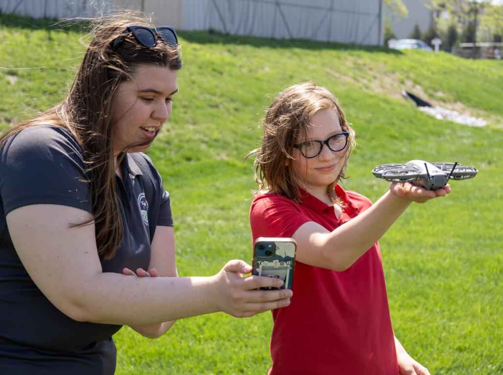 A college student assists a child who is holding a small drone in their hand. They are outdoors on a sunny day.