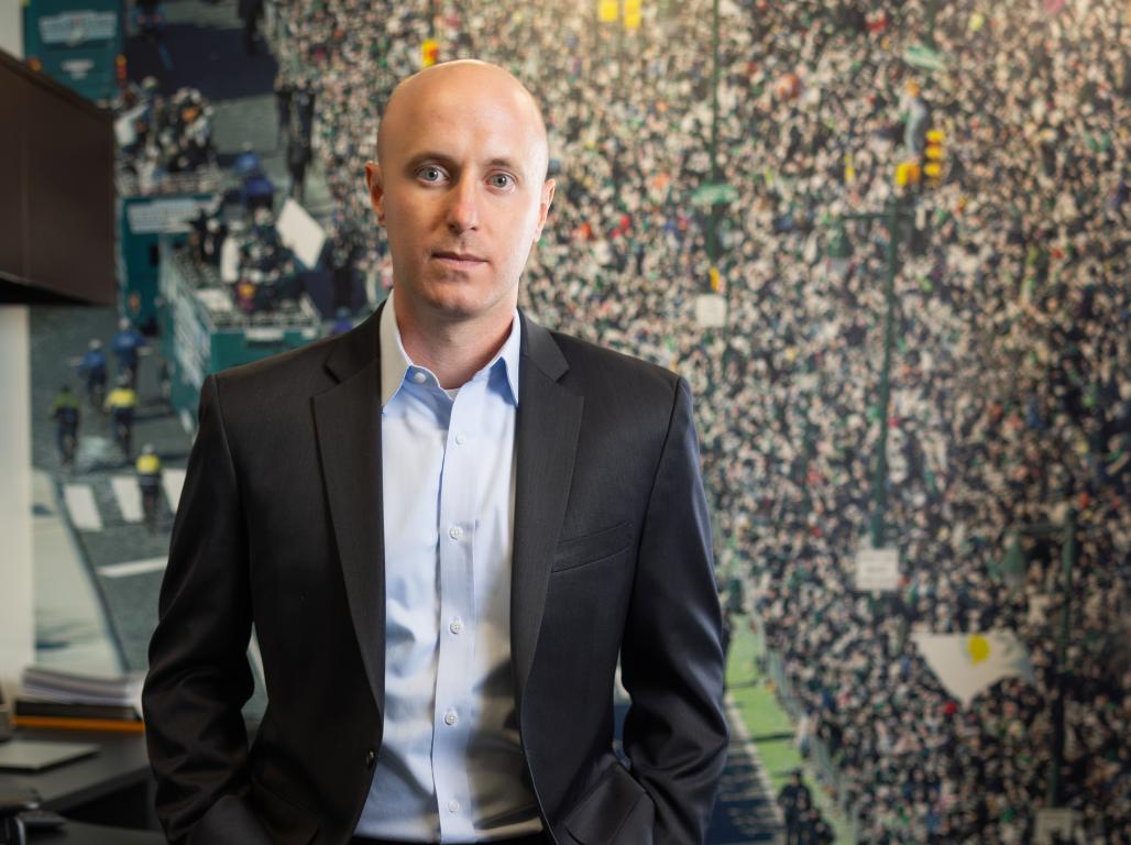 Professional portrait of Bryce Johnston, VP with the Philadelphia Eagles, leaning against a desk with hands in suit pockets.