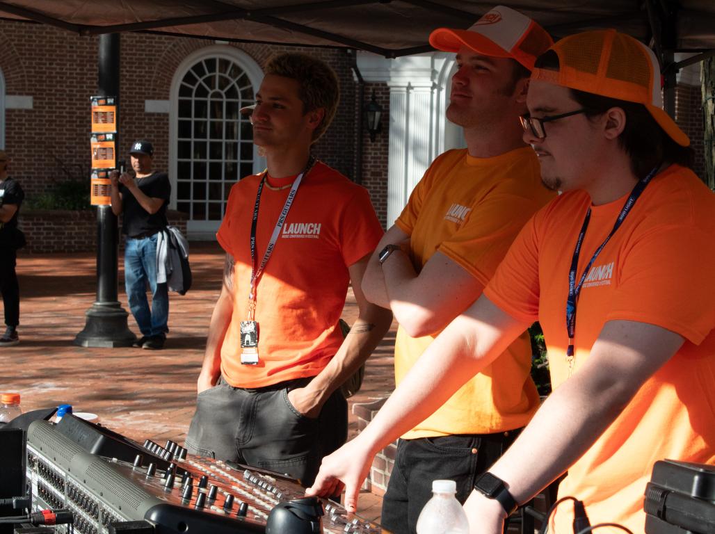 Three people wearing orange shirts and ID badges stand over an audio mixing board. A brick building is visible in the background.