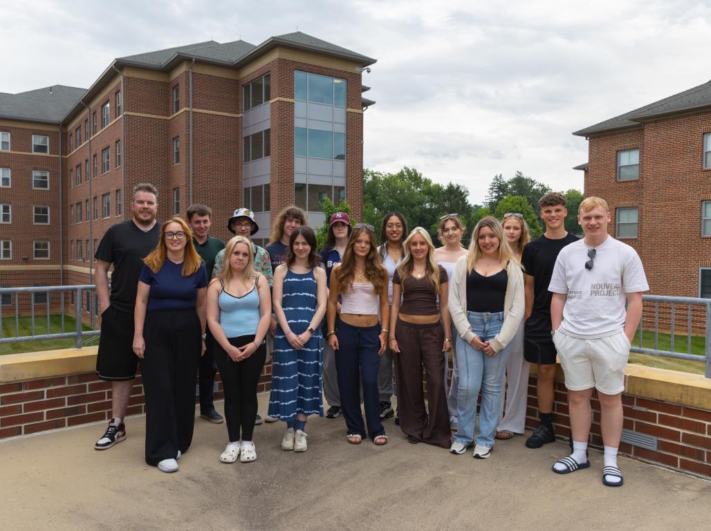 A group of 16 visiting students and two chaperones from York College & University Centre in England pose together on an outdoor terrace at York College of Pennsylvania, with brick campus buildings and trees in the background.