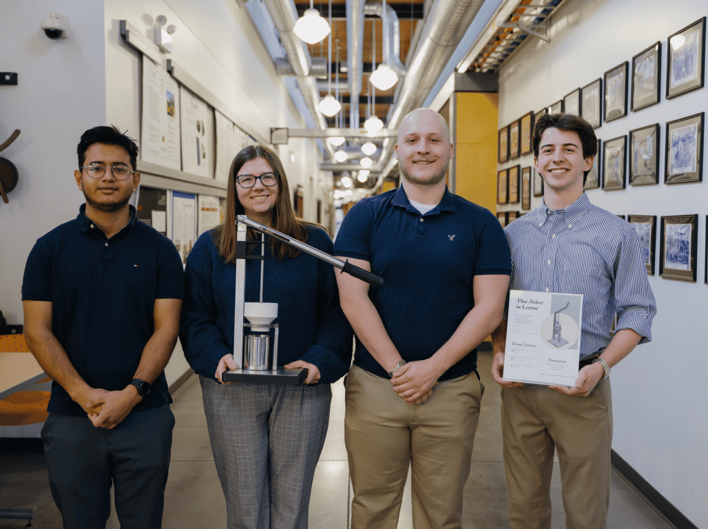 Four York College Mechanical Engineering students stand with their completed class project, with Aidan Heck ’27 at right holding the project poster.