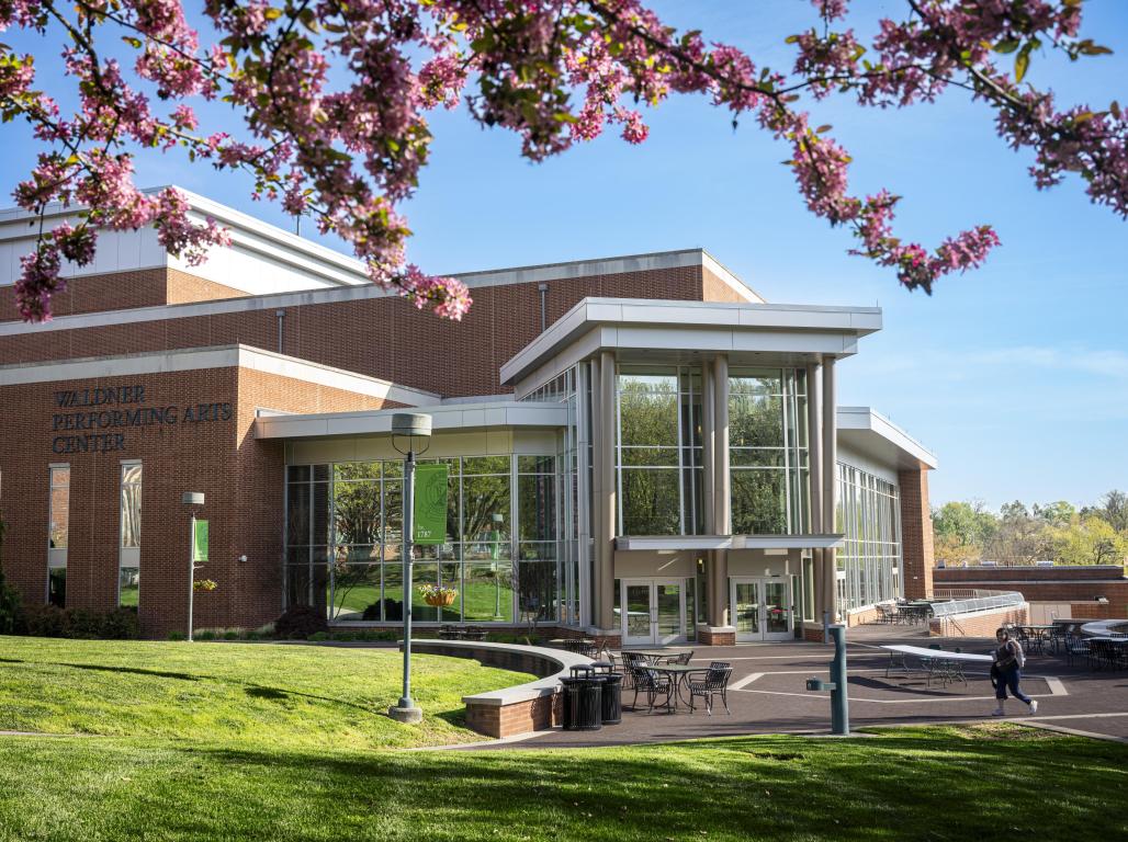 A spring photo of the WPAC building. A large brick building with a glass front. Green lawn in front with blue skies and a flowering tree branch at the top of the photo.