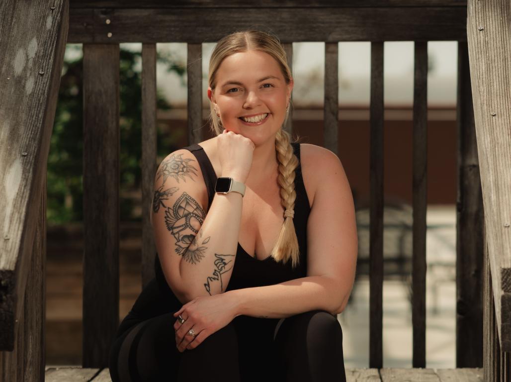 Kirsten Blair-Johnson ’18 smiles while sitting on wooden steps outdoors in yoga attire.