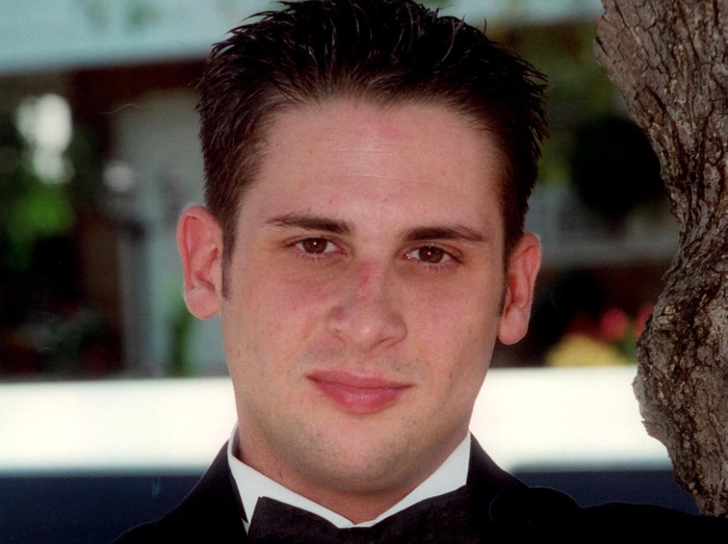 A man in a black tuxedo and bow tie poses outdoors with his arms crossed beside a tree.