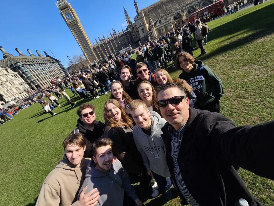 A professor takes a selfie with a group of students in front of a clock tower.