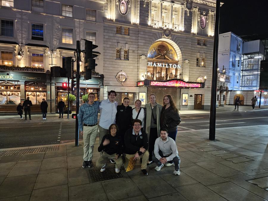 A group of individuals pose in front of a theater marquee with the words "Victoria Palace" and "Hamilton" in lights.