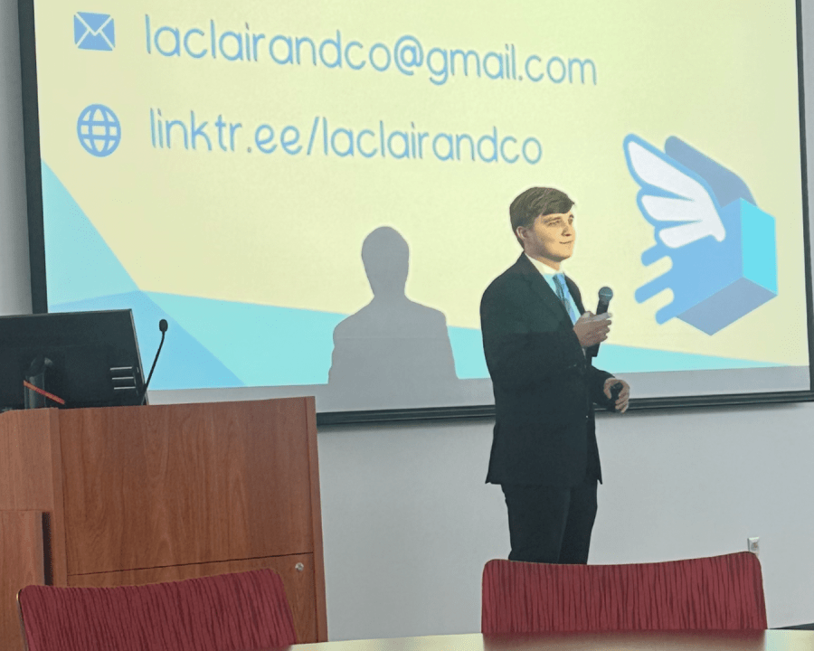 Person in a suit and tie speaking into a microphone in front of a projector screen with contact info afor