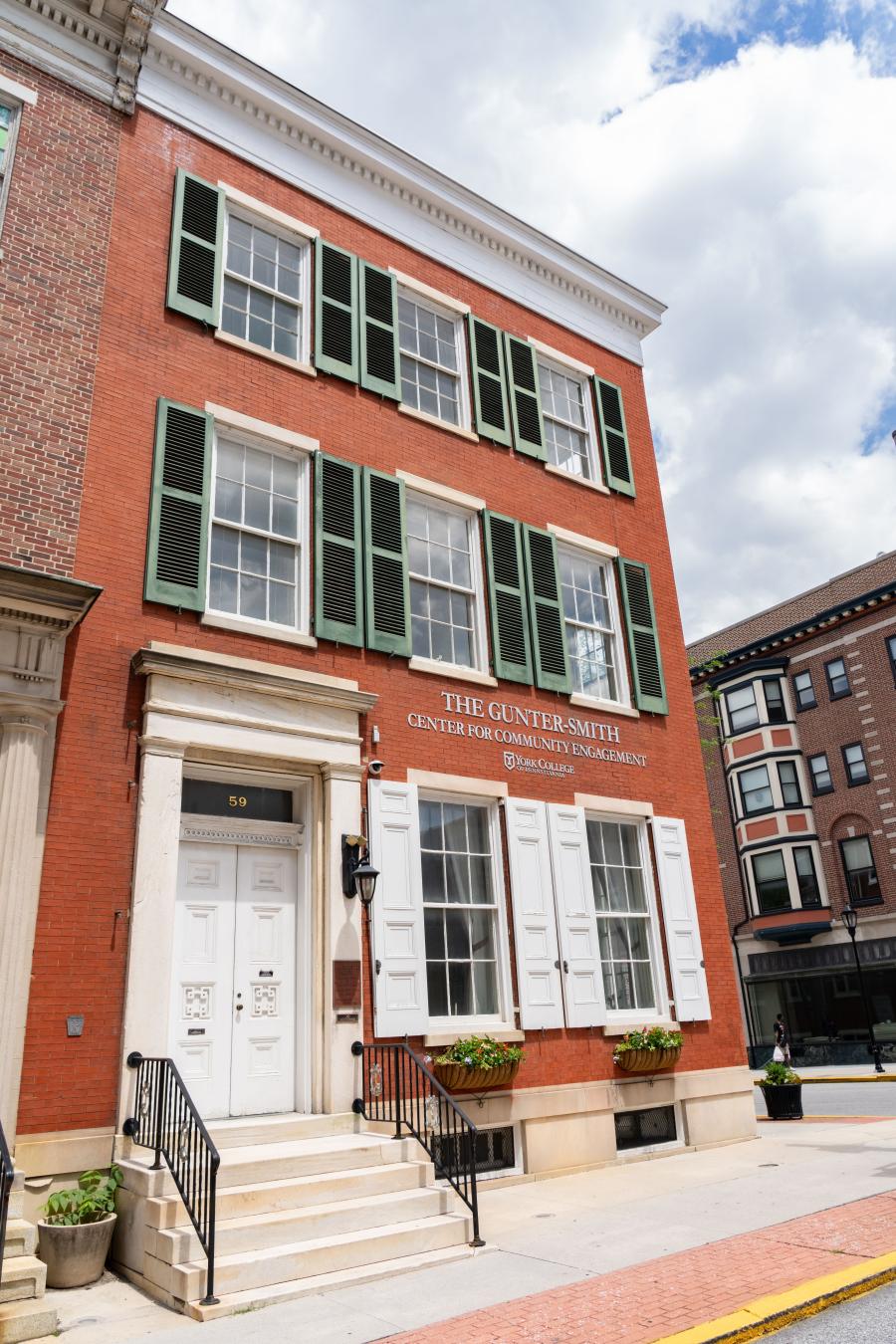 Historic brick building with a white door and green shutters and the words "Gunter-Smith Center for Community Engagement" in silver lettering above a York College logo. Corner of a city block on a sunny day.