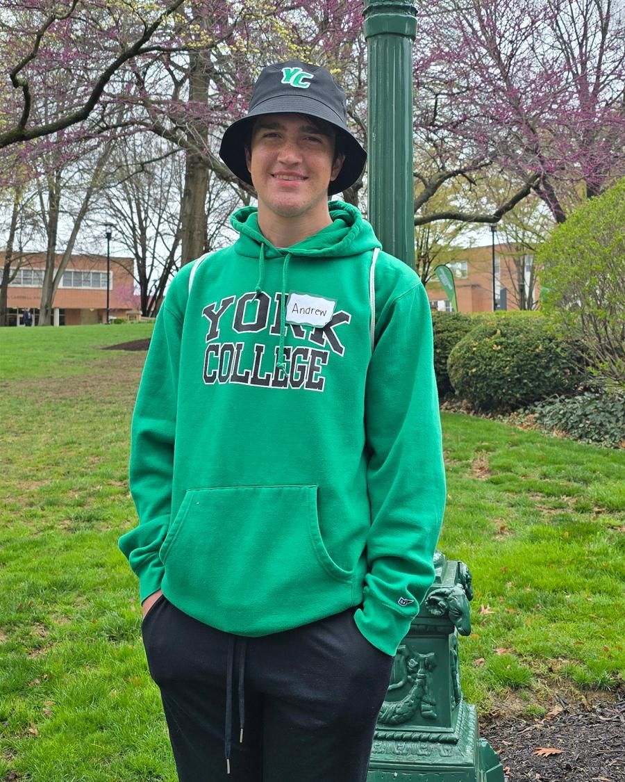 Person wearing a green York College sweatshirt smiles with springtime trees in the background.