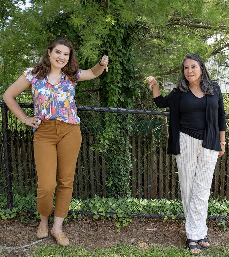 Professors AnaLu MacVean, Environmental Horticulture, Emily Rund, Filmmaking, stand side by side in front of an invasive plant making the thumbs down hand motion