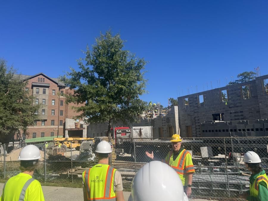 A person wearing protective gear gestures toward a construction site while a group listens in the foreground.