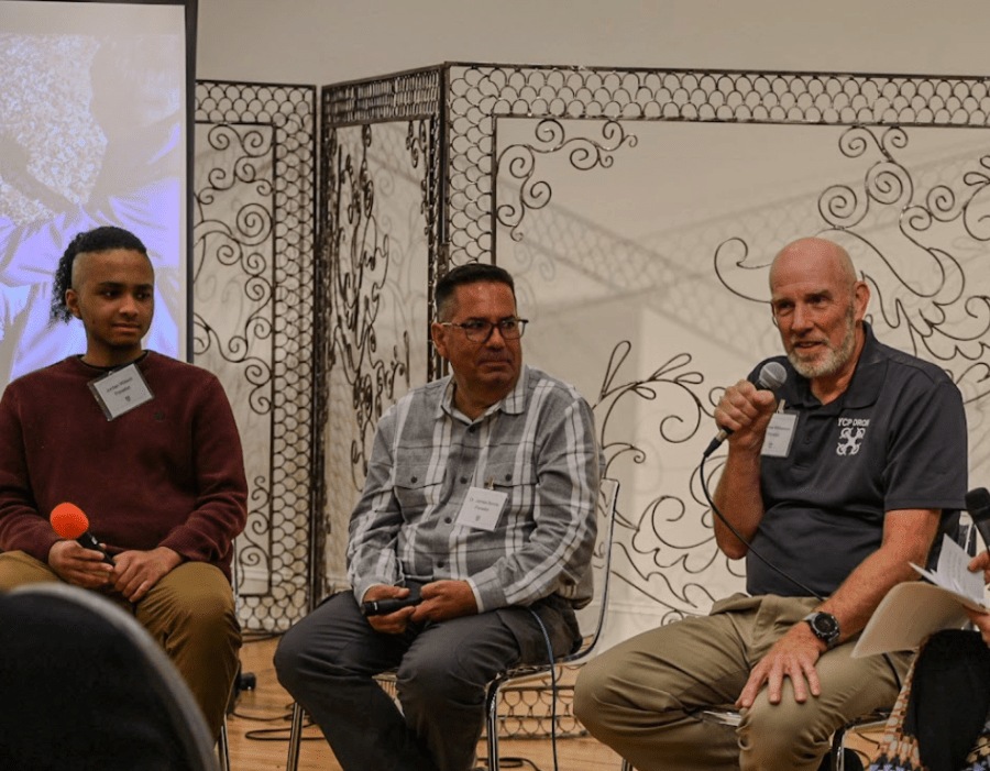 Professor Wilkerson hold a microphone as he speaks to an audience with two people beside him