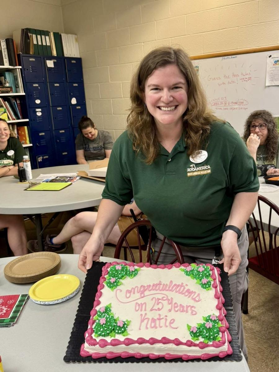 Katie Govern wearing a ZooAmerica uniform shirt while posing with a cake decorated with "Congratulations on 25 Years Katie" in a break room.