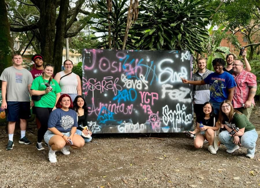 A group of people pose with a large spray-painted board. They appear to be in a tropical setting.