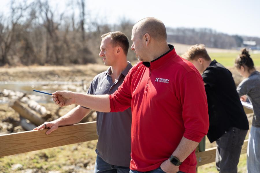 Person in a red Kinsley Construction shirt pointing a pen at a muddy field while explaining something to someone beside them. Two other people can be seen taking notes behind them.
