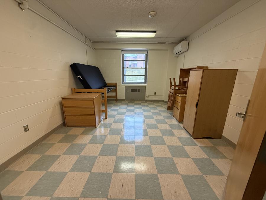 Empty bedroom in Codorus Hall. Single bed, dresser, side table, and desk. Window on the fall wall with air conditioner unti on side wall.