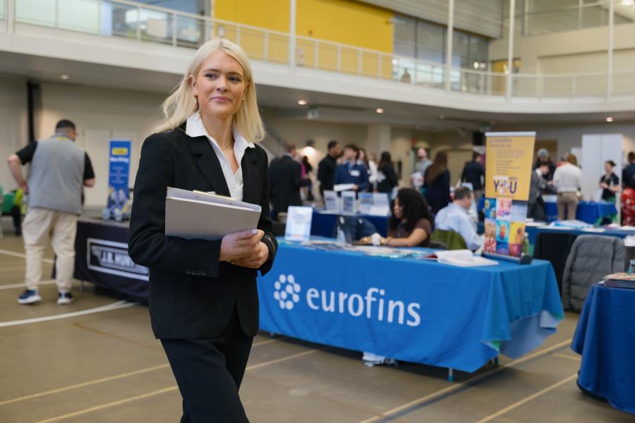 College student in business attire walks through a professional networking event, holding a folder.