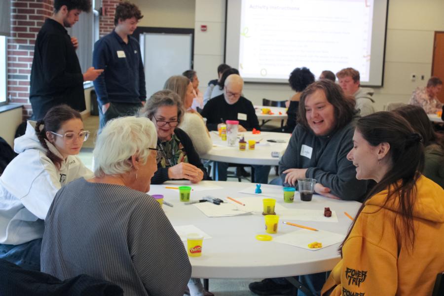 A group of people of varied ages sit around a round table. They appear to be engaged in a creative activity.