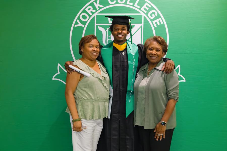 A college graduate in regalia stands with loved ones on either side. A green York College backdrop is visible behind them.