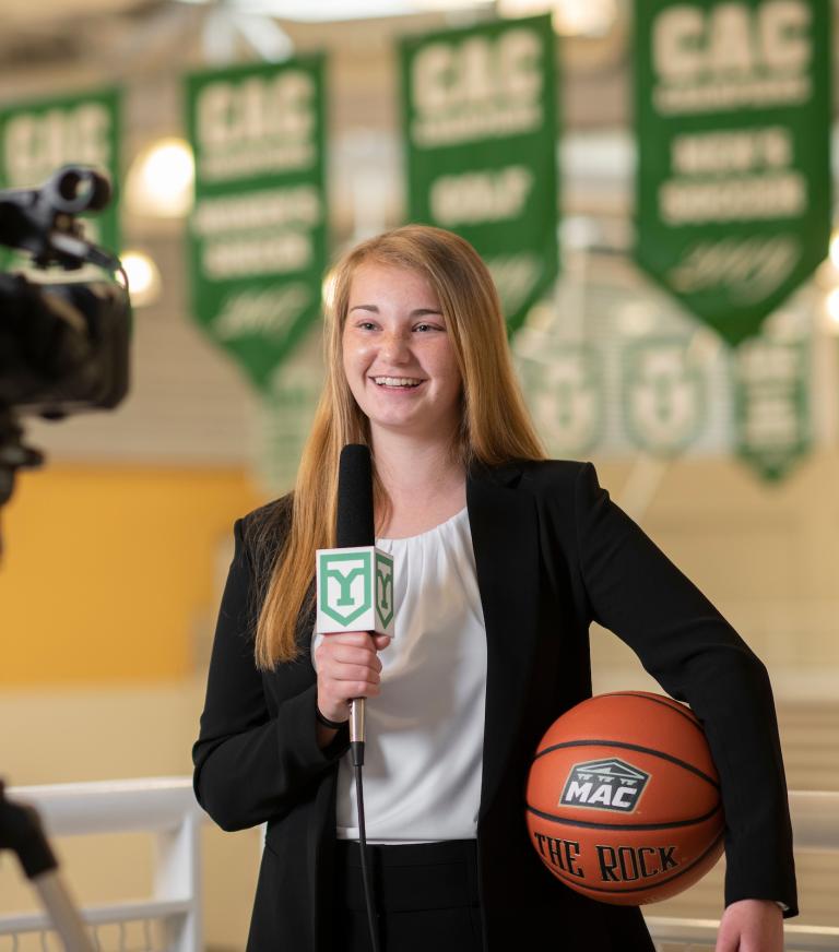 A student holding a basketball with the left arm holds a microphone with the right hand and faces a video camera.