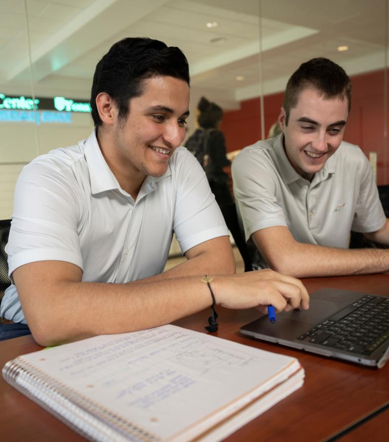Two students are smiling looking at information on a computer in the NASDAQ Training Lab