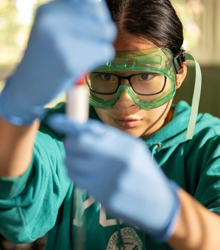 A student wearing safety goggles and lab gloves stares intently as they handle a test tube in a chemistry lab.
