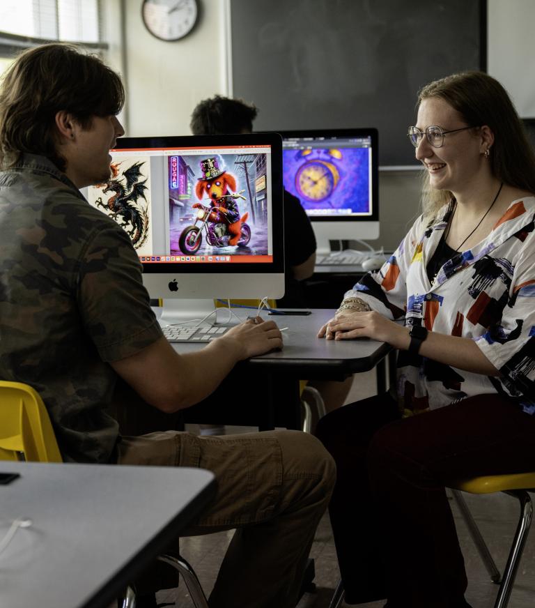 Two individuals engaged in conversation in a classroom setting with AI art visible on various computer screens.