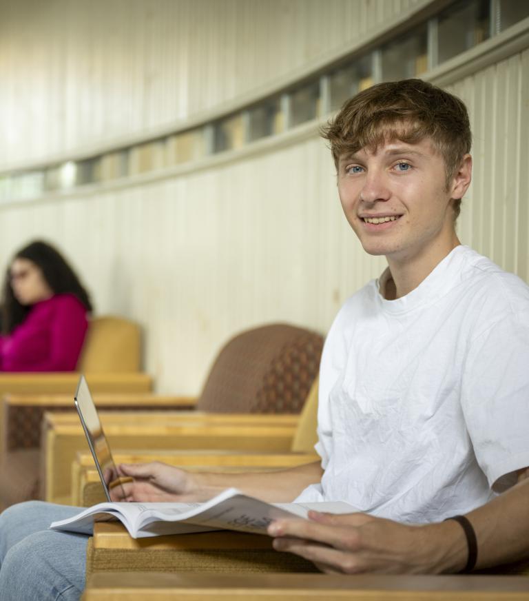 College student wearing a white tee shirt sits in a chair in a hallway, holding a book and laptop while smiling at the camera. Another person in a pink shirt is visible in a chair in the background.