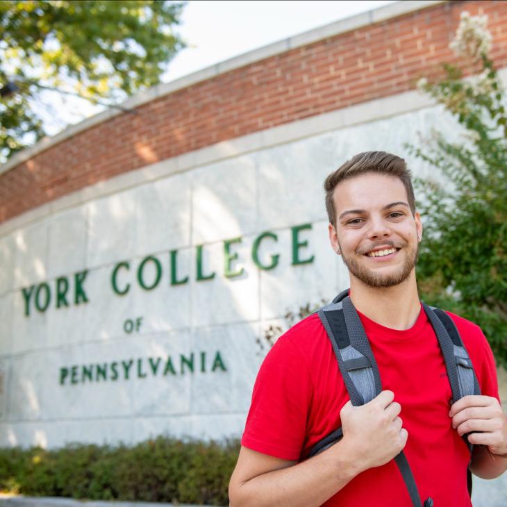 Student with backpack smiles while standing in front of York College entrance signage
