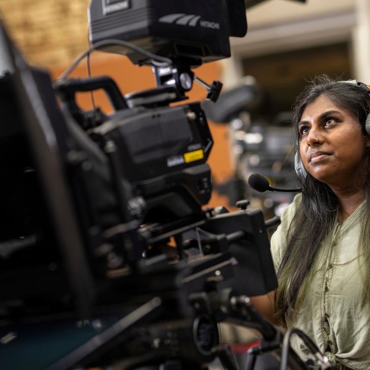 A student seated behind a video camera looks at the preview screen.