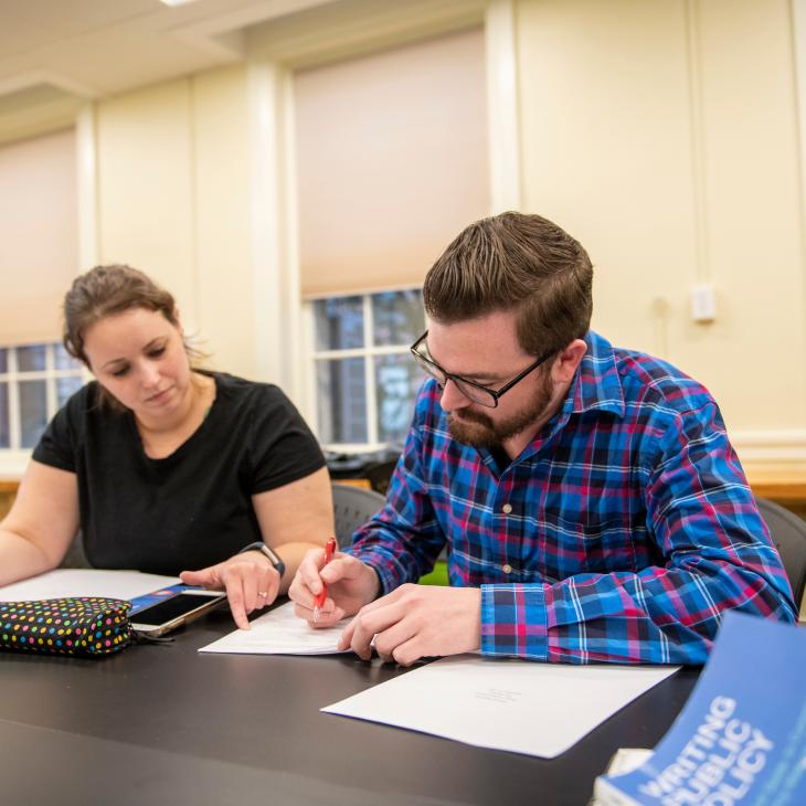 Two students look at papers spread over a table. One is writing while the other holds a binder.