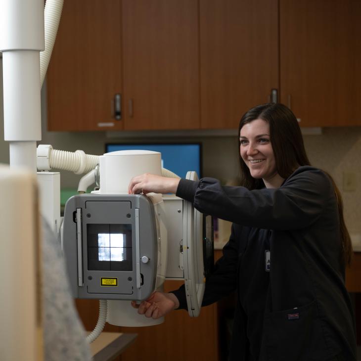 A student wearing scrubs adjusts a large imaging machine to x-ray a patient in a hospital lab. 