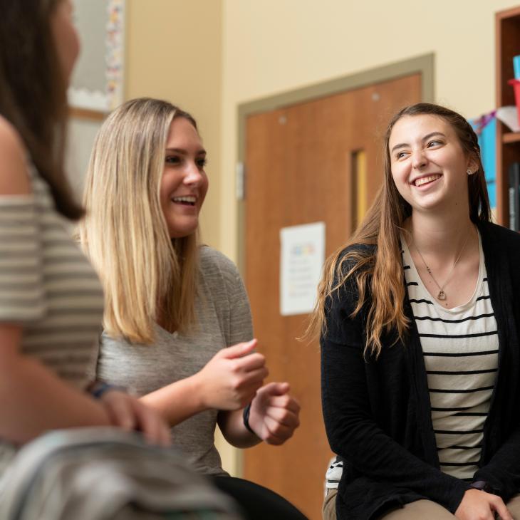 Three students gather in a classroom, chatting and smiling as books and supplies are visible on shelves in the background.