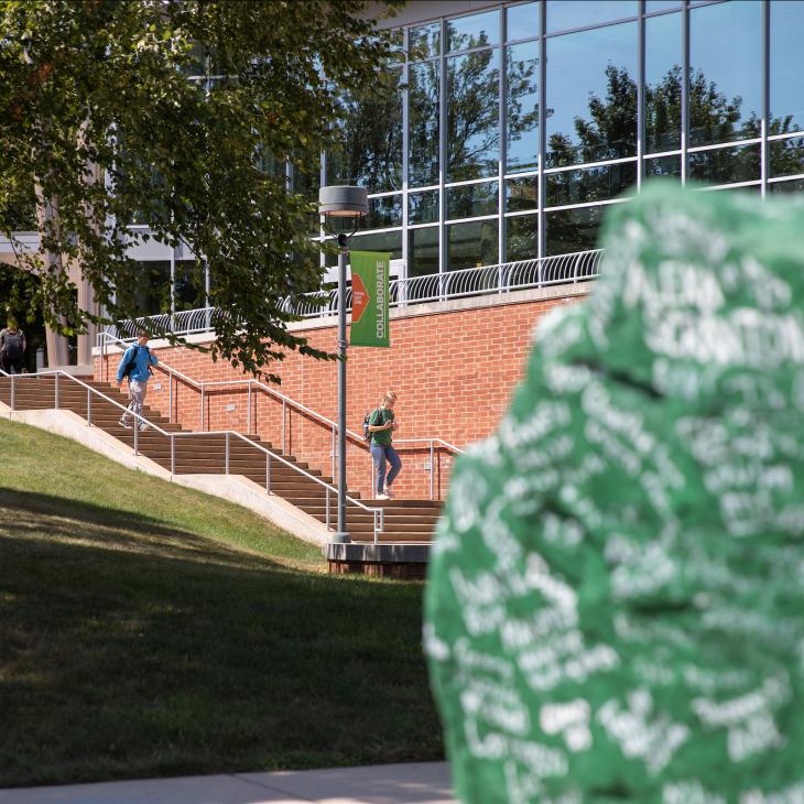 Students walk down outdoor steps beside the Performing Arts Center building. In the foreground, Ol Spart (a green boulder covered in white painted signatures) is visible.