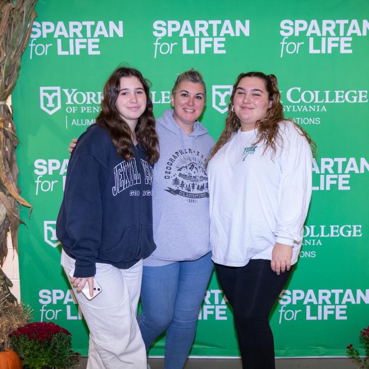 Three people stand in front of a backdrop reading "Spartans for Life" surrounded by pumpkins and cornstalks and other fall decor.