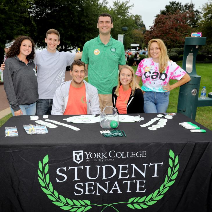 Six members of the Student Senate, dressed in colorful shirts, pose behind the Student Senate table at the Fall Fest. On the table an assortment of souvenirs - pens, buttons, water bottles and others - are laid out for visitors.