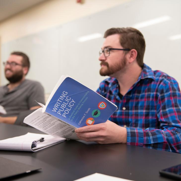 Graduate student in a blue plaid shirt listens to instructor, referencing their "Writing Public Policy" book.