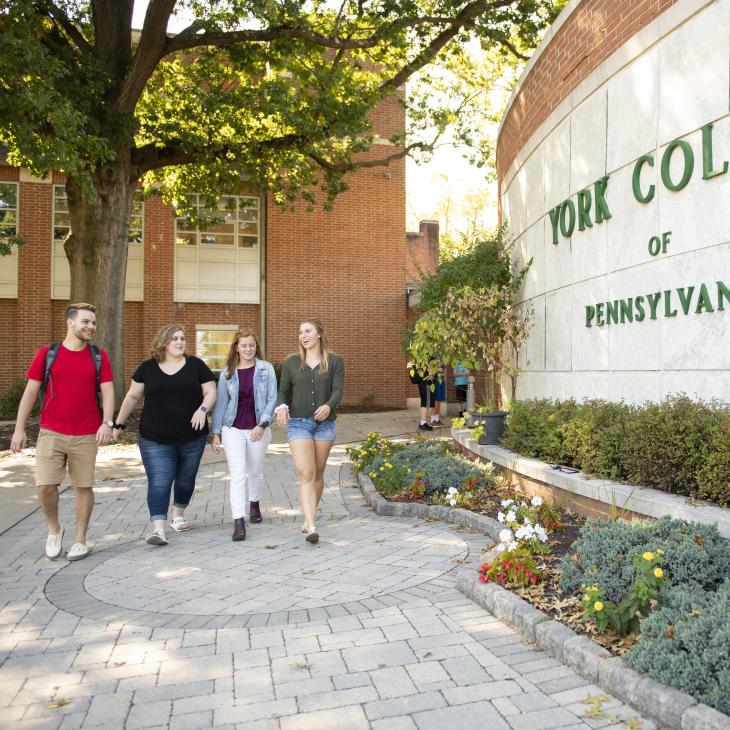 4 Students are walking from Wolf Hall in front of a York College sign