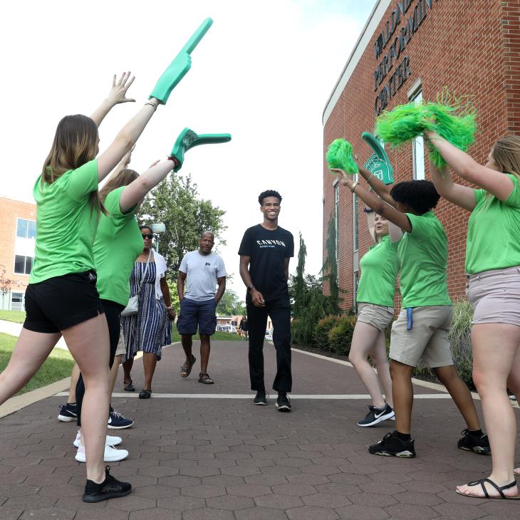 A group of student orientation leaders wearing green t-shirts wave green pom poms and cheer as a new student approaches on campus.